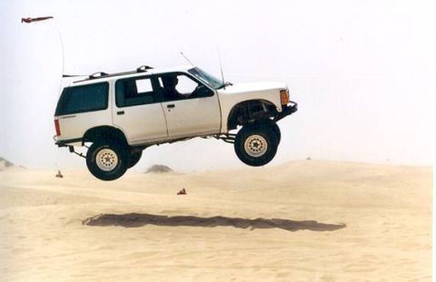 1985 Ford Bronco II - A white SUV is captured mid-air while jumping over sand dunes.