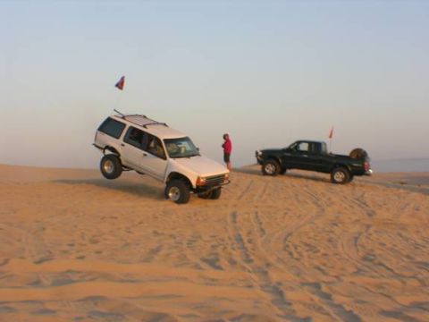1985 Ford Bronco II - Two off-road vehicles navigate a sandy landscape under a clear sky.