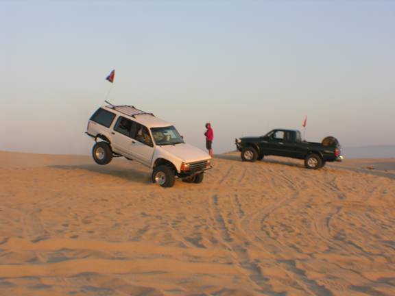 White SUV airborne over sand dune, black pickup truck on dune crest
