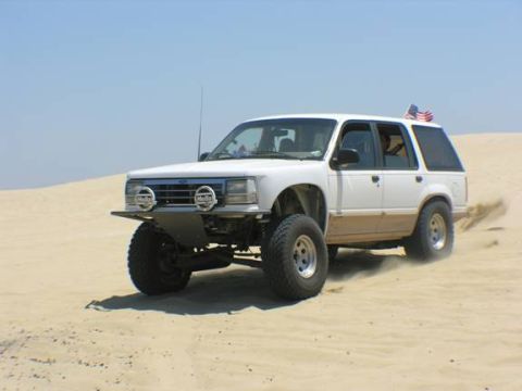 1985 Ford Bronco II - A white SUV is shown driving on a sandy terrain, with an American flag attached to its roof.