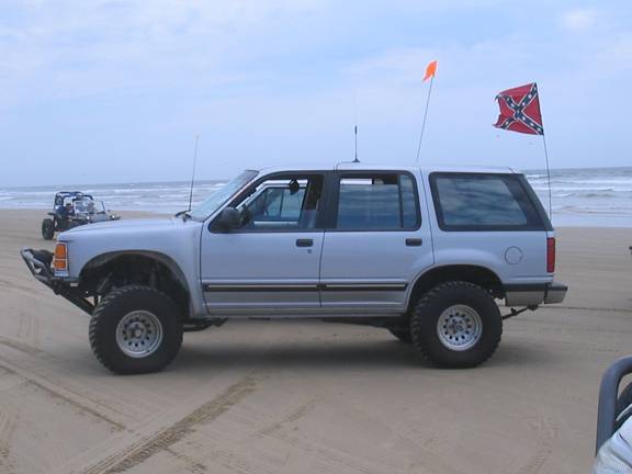 Silver SUV with off-road tires on a beach, another vehicle in background. Flags on poles