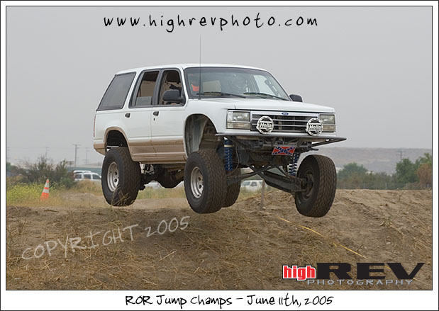 White SUV with lifted suspension mid-air over a dirt mound. It has large off-road tires and auxiliary lights on the front bumper