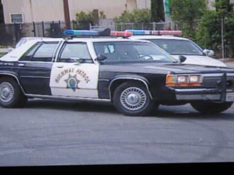 1991 Ford LTD Crown Victoria - photographed television frame showing two black-and-white Highway Patrol sedans with lightbars parked side by side near a fence.