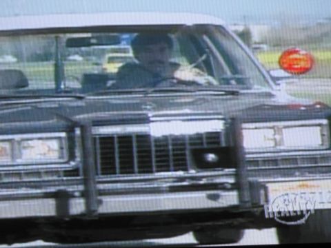 1991 Ford LTD Crown Victoria - photographed television frame showing a mustached driver inside a dark full-size sedan with a chrome grille and front brush guard.