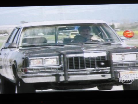 1991 Ford LTD Crown Victoria - photographed television frame showing a mustached driver in a black-and-white full-size sedan with chrome grille and a Fox Reality logo.