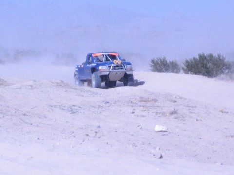 Blue off-road race truck cresting a distant sandy ridge in heavy desert dust