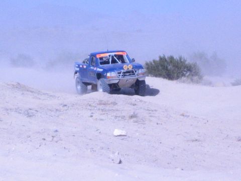Blue off-road race truck with front lights cresting a sandy desert berm