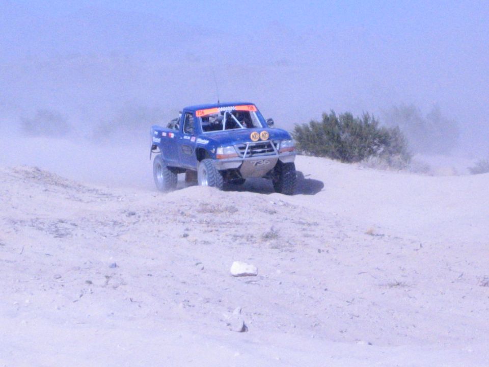 Blue off-road race truck with front lights cresting a sandy desert berm