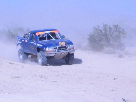Blue off-road race truck with yellow KC lights driving on a sandy desert track