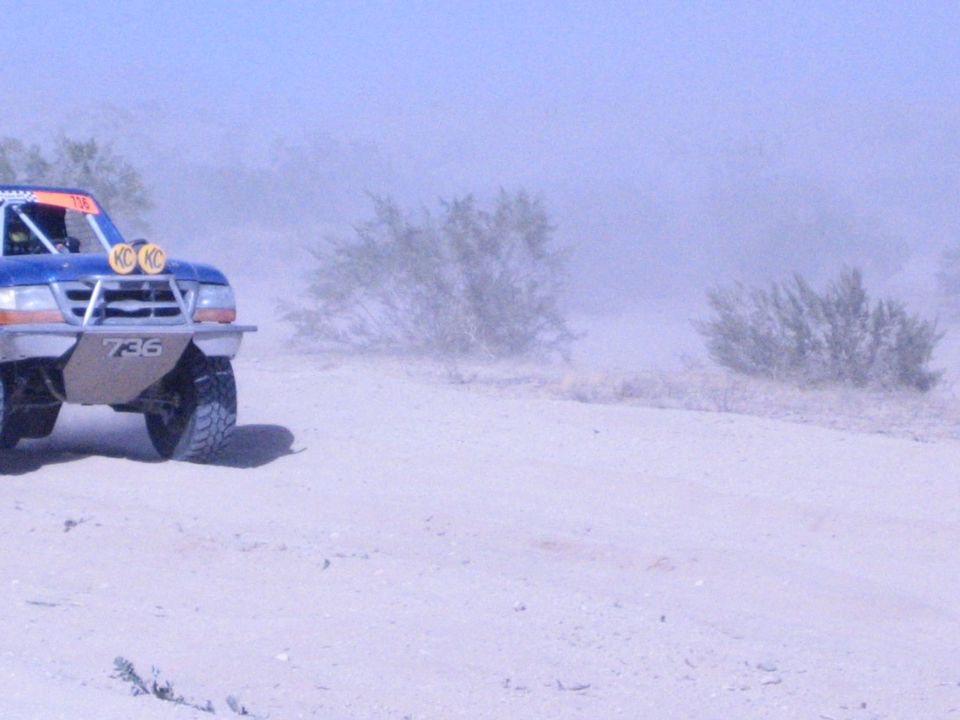 Front corner of blue off-road race truck 736 with yellow lights in desert dust
