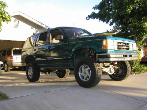 1984 Ford Bronco - This lifted green SUV is shown from the front 3/4 angle, with another SUV visible in the background.