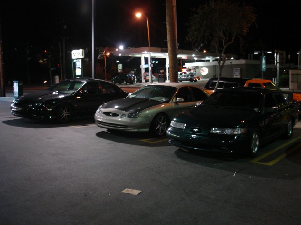 Three sedans parked at night, one black, one silver with a dark hood, and one dark green. Gas station in background