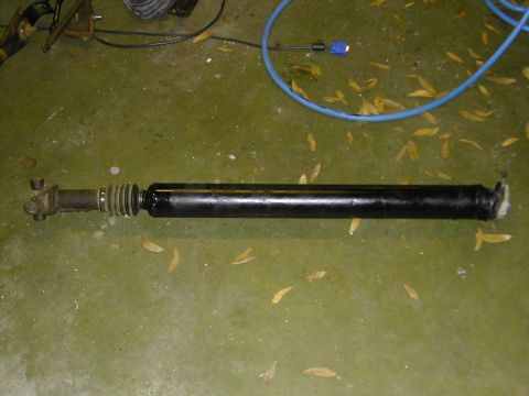 1993 Ford Bronco - A removed slip-yoke driveshaft laid out on a green-painted concrete garage floor, showing the black-painted tube, U-joint yoke and a ribbed…