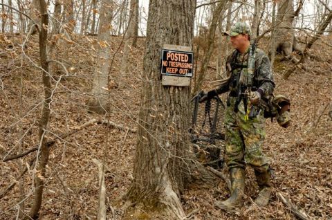 1986 Ford Bronco - A hunter in full camouflage and rubber boots carries a climbing treestand through leaf-covered winter woods, passing a tree nailed with a r…