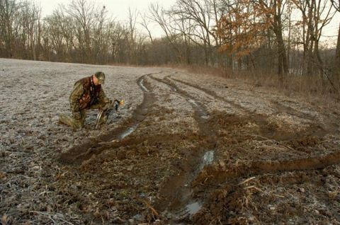 1986 Ford Bronco - A person in camouflage kneels in a frosty muddy field measuring deep water-filled tire ruts that curve across the ground toward bare winter…