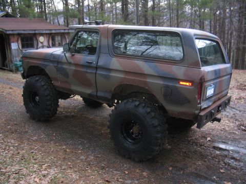 1979 Ford Bronco - This lifted Ford Bronco, painted in camouflage, sits parked on a dirt road next to a rustic cabin.