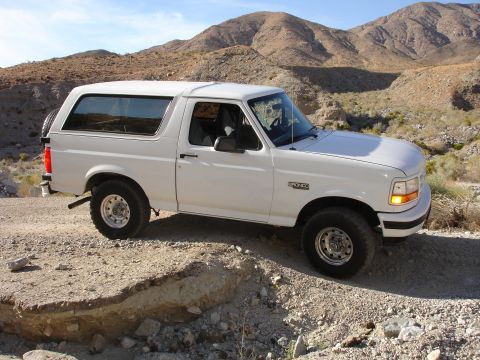 1996 Ford Bronco - A white 1996 Ford Bronco is seen from a three-quarter front view on a dusty, rocky terrain with mountains in the background.