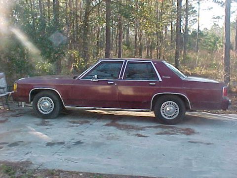 1994 Ford Crown Victoria - Maroon sedan with chrome trim and wire wheel covers is parked on a concrete surface with trees in the background.