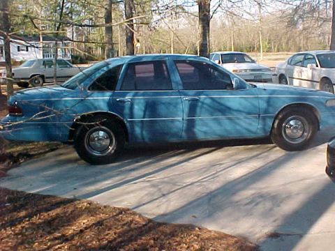 1994 Ford Crown Victoria - A blue 1994 Ford Crown Victoria is seen from the side, parked on a concrete driveway with other vehicles in the background.