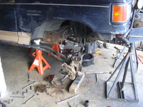 1991 Ford Bronco - The front suspension of a Ford Bronco is disassembled and supported by jack stands, with tools and fluids visible on the garage floor.