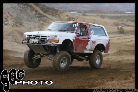 1984 Ford Bronco - This modified Ford Bronco is captured mid-jump during an off-road race, its lifted suspension and racing modifications.