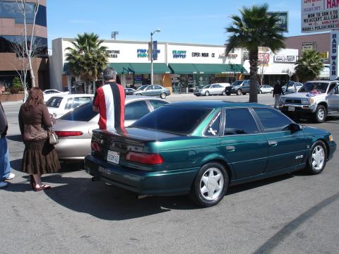 1993 Ford Taurus - A green sedan is parked on a street with several other vehicles and businesses visible in the background.