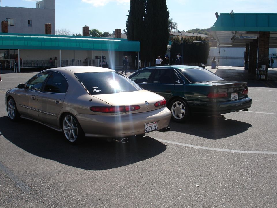 Gold sedan and green sedan parked side-by-side, rear view. Gold sedan has a spoiler and dual exhaust