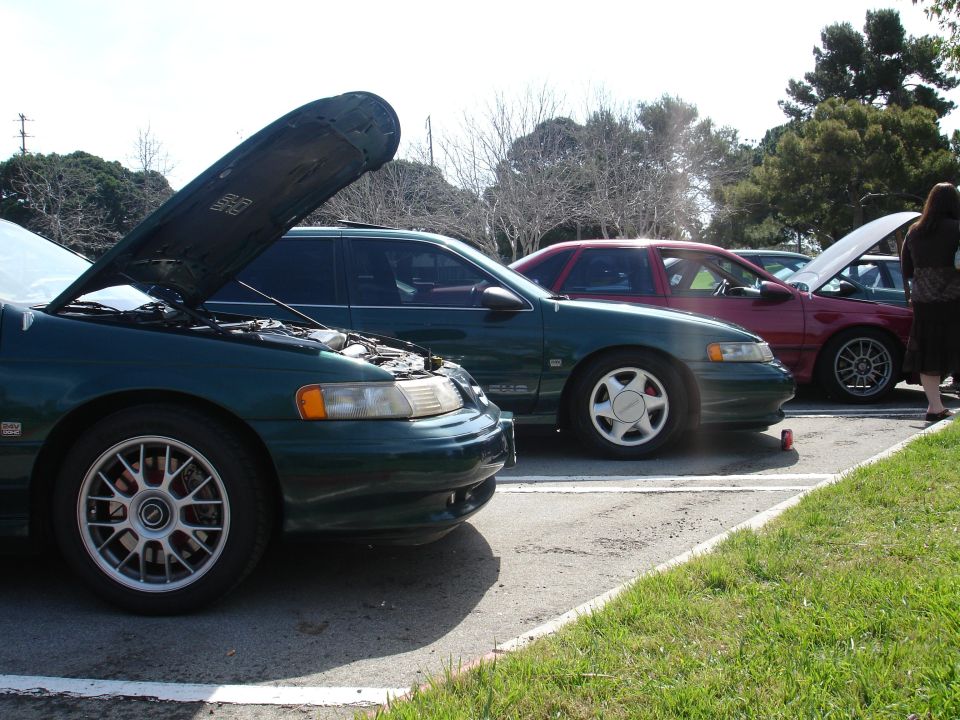 Three cars parked in a lot, two with hoods open. The front car is dark green, the middle is dark green, and the rear is red