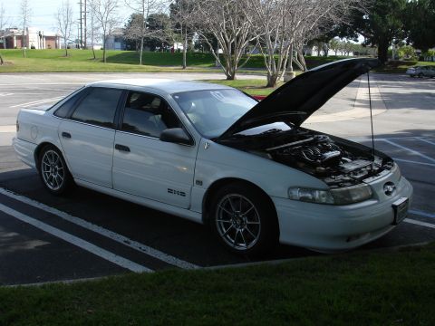 1993 Ford Taurus - A white sedan is parked with its hood open, revealing the engine compartment and aftermarket wheels.