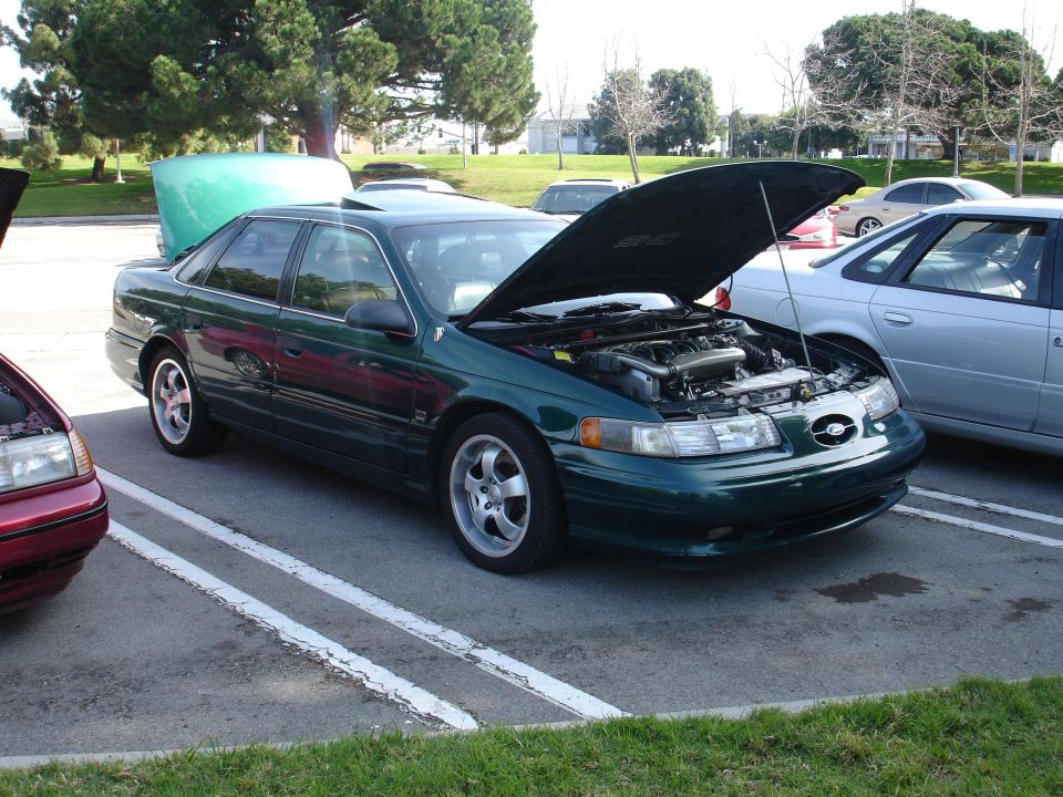 Green sedan parked in lot, hood open showing engine. Another car to the left also has hood open