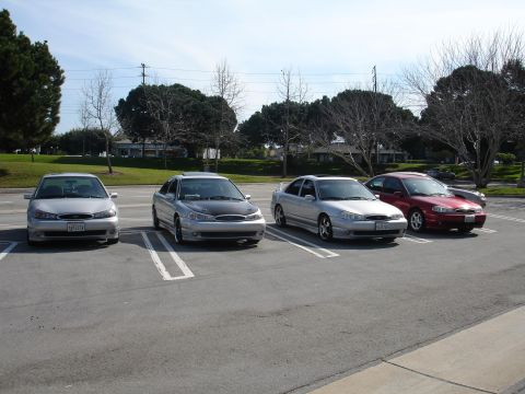 1993 Ford Taurus - Four sedans are parked in a lot on a sunny day. Three are silver, one is red.