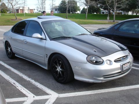 1993 Ford Taurus - This sedan features a contrasting black hood and aftermarket wheels, parked alongside another vehicle.