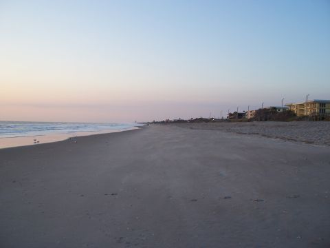 1994 Ford Probe - A wide expanse of beach stretches towards the ocean, with buildings visible along the coastline under a soft, pastel sky.