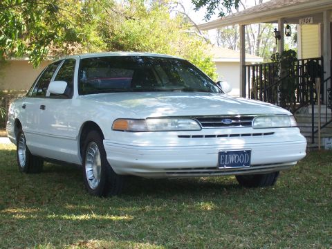 1997 Ford Crown Victoria - A white Ford Crown Victoria is parked on a grassy lawn in front of a house with a visible license plate that reads 'ELWOOD'.