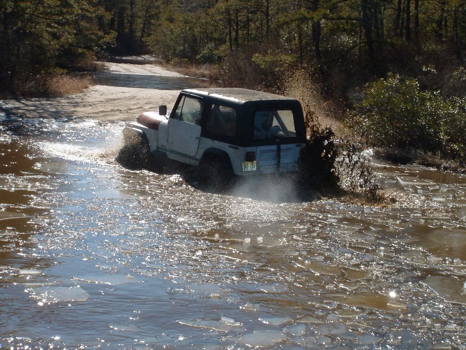 White soft-top Jeep Wrangler throwing spray sideways through a wide flooded sand trail