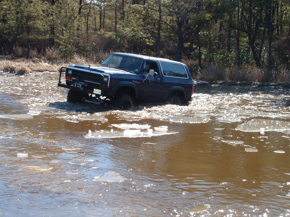 Dark Ramcharger-style SUV with brush guard plowing a brown icy flood, ice chunks floating around it