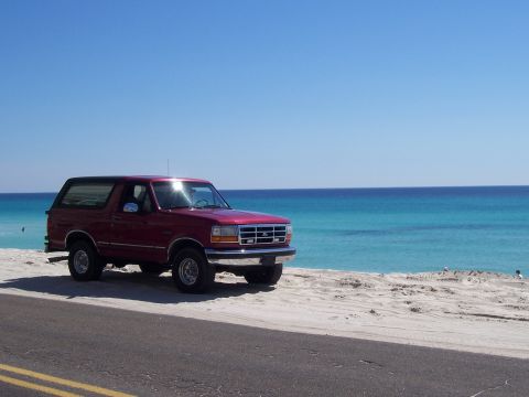 1995 Ford Bronco - Three-quarter front view of a red full-size Ford Bronco with a black removable hardtop, parked on white sand above a roadway with turquoise…