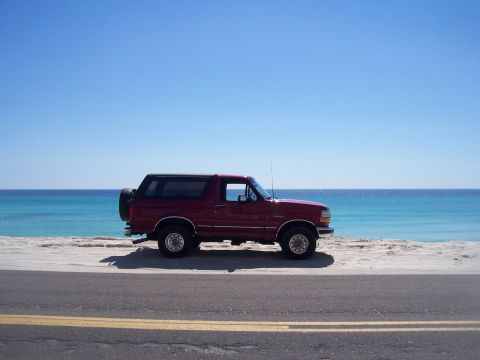 1995 Ford Bronco - Passenger-side profile of a red full-size Ford Bronco with a black removable hardtop, parked on white sand beside a coastal road with turqu…