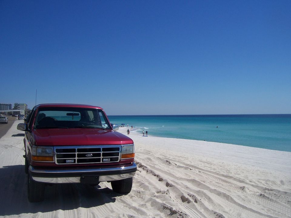 Red 1990s Ford truck parked on white beach sand by turquoise ocean