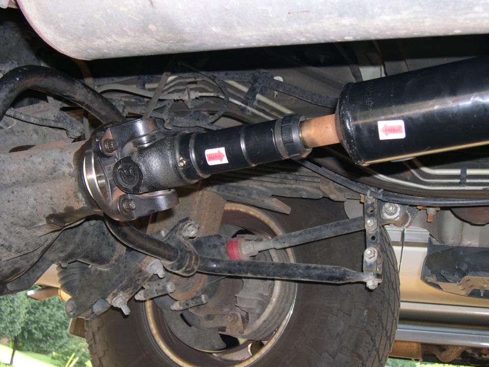 Underside view of rear driveshaft U-joint at axle pinion, with leaf springs and rear tire visible.