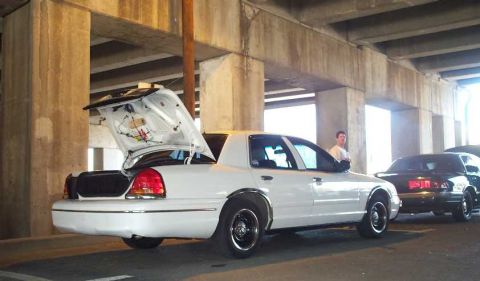 1998 Ford Crown Victoria - A white 1998 Ford Crown Victoria is seen from the rear with its trunk open, parked under an overpass next to another vehicle.