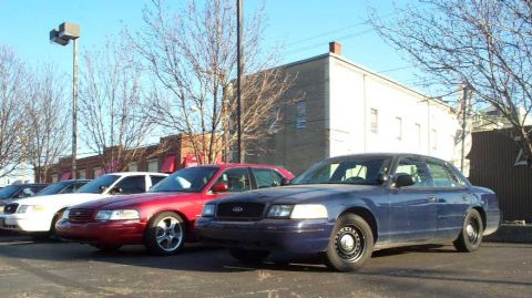 1998 Ford Crown Victoria - A blue 1998 Ford Crown Victoria is parked in a lot alongside other sedans under a bright blue sky.