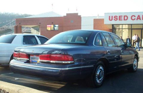 1998 Ford Crown Victoria - A dark blue sedan is parked in front of a building with a 'USED CAR' sign, with another white car beside it.