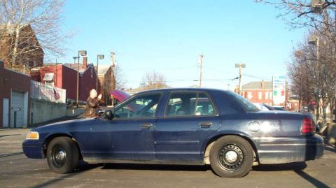 1998 Ford Crown Victoria - A person stands by a dark blue sedan with its hood open on a sunny day.