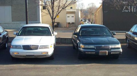 1998 Ford Crown Victoria - Two sedans are parked in a lot, with a building and trees in the background.