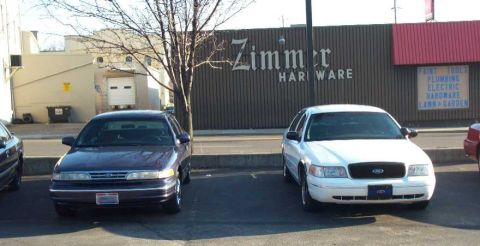1998 Ford Crown Victoria - Two Ford Crown Victorias, one dark purple and one white, are parked in front of Zimmer Hardware.