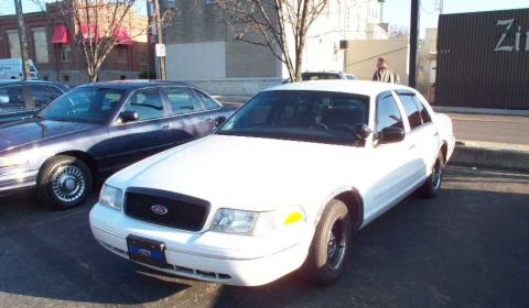 1998 Ford Crown Victoria - A white police car is parked in a lot next to a dark sedan, with buildings in the background.