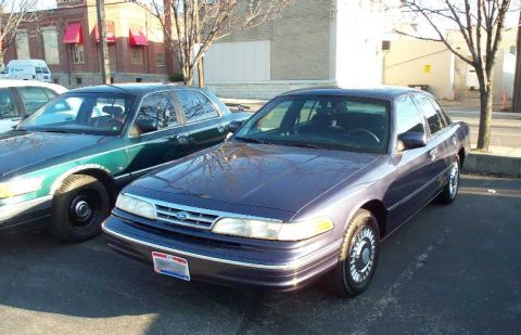 1998 Ford Crown Victoria - A dark purple Ford Crown Victoria is parked next to a green car, with buildings in the background.
