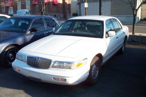 1998 Ford Crown Victoria - A white Mercury Grand Marquis is parked in front of a building, with another car next to it.
