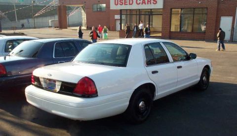 1998 Ford Crown Victoria - A white sedan is parked at a used car lot with other vehicles and people in the background.